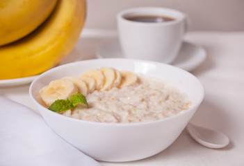 Oatmeal porridge with banana in the white bowl, cup of coffee on the white table. Health breakfast.