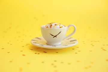 Ceramic white cup with glitter stars inside and small saucer with clock face icon. Yellow background.