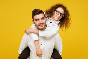 Portrait of happy young brunette woman in eyeglasses enjoying piggyback ride on boyfriend's back against blank yellow studio wall background. Love, romance, togetherness and happiness concept