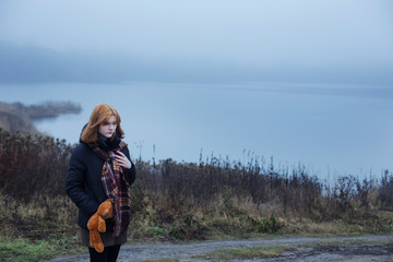 Sad teenager girl with  teddy bear on country road by foggy  lake.  Concept of adolescence and adolescent problems.