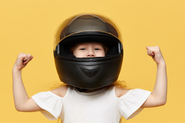 Isolated shot of little girl racer posing against yellow studio wall background wearing black safety motorcycle helmet demonstrating her bicep muscles. People, extreme sports and adrenaline concept