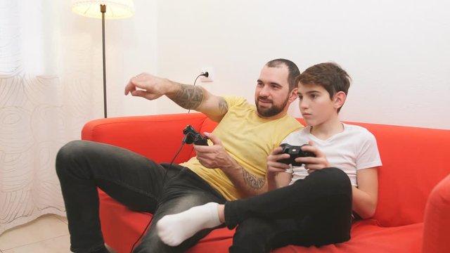 Caucasian family, positive smiling man and happy kid boy sit on sofa and play video games together, holding joystick in hands