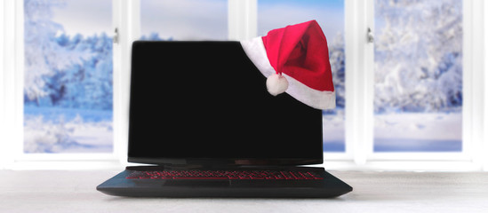 Laptop and gifts on the table with Santa Claus hat at home with a panoramic view through a window of snowy trees in a winter forest, the concept of a holiday and preparation for Christmas