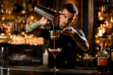 Professional male bartender pouring a alcoholic drink from the steel shaker to the glass through...