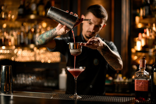 Professional male bartender pouring a alcoholic drink from the steel shaker to the glass through the sieve