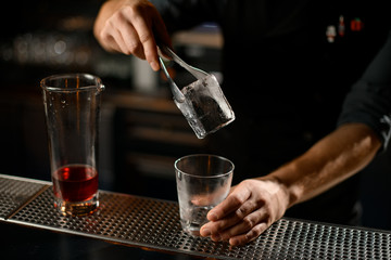 Obraz na płótnie Canvas Male bartender putting an ice cube with tweezers to the empty cocktail glass