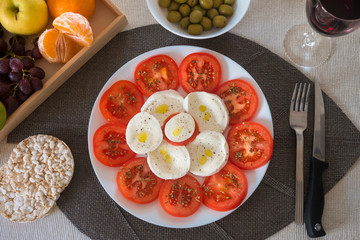 Dieting with a light and healthy dish of caprese salad with tomatoes and mozzarella. Accompanied by rice cakes, green olives and fruit. A glass of red wine