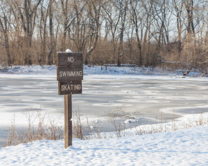 Wooden no swimming, ice skating sign with snow covered ice on frozen lake in background. Concept of winter safety and danger of thin ice