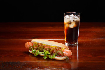 Hot dog with mustard, lettuce and jalapenos on a wooden table. Nearby is a glass of cola with ice.