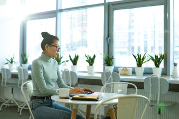 Serious female student in casualwear sitting by table in front of laptop in cafe