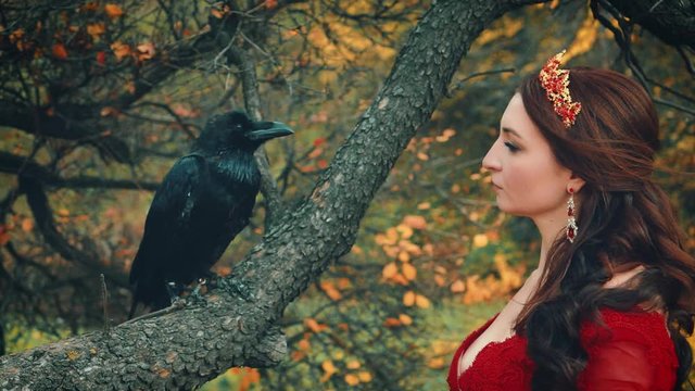 Close-up portrait of queen stroking touching big raven with tenderness. wild bird sits on branch of autumn tree with yellow leaves. Hair flying in wind. Gold crown jewelry. Backdrop fabulous nature