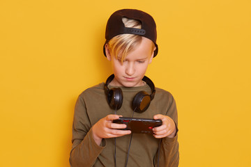 Child playing online games, little boy with digital device isolated over yellow background, male child wears green shirt and cap with headphones around neck, kid looks concentrated. Technology concept
