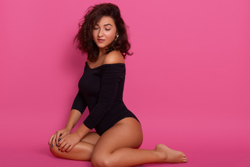 Studio shot of mysterious woman wearing black combi dress, sitting on floor, looking down, girl with dark wavy hair, lady posing isolated over pink background, gitl model with slim perfect legs.