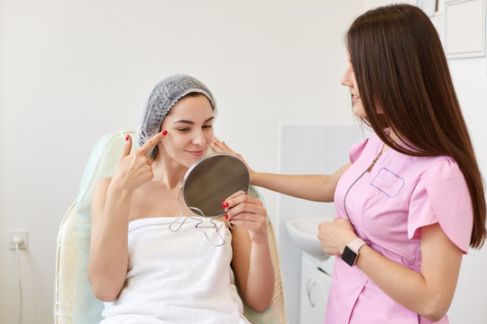Image of beautiful delighted client looking at reflection in mirror, checking skin condition after beauty procedure, lying at cosmetologic chair, consulting helpful cosmetologist in pink lab coat.