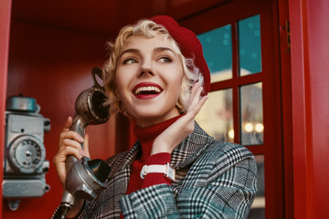 Fashionable happy smiling blonde woman wearing red beret, turtleneck, checkered coat, white wrist watch, talking on the retro phone, posing in red  call box. Copy, empty space for text - obrazy, fototapety, plakaty