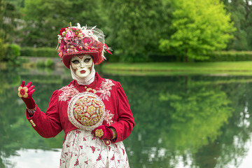 A woman standing in mask and red masquerade costume agains the pond background - 311239074