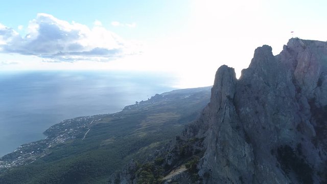 Aerial view of sharp rocky cliffs above blue ocean on cloudy sky background. Shot. Flying over the mountain peaks and coastal small city.