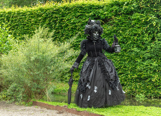 A woman standing in mask and black masquerade costume with a lamp against the pond backgroun - 311240042