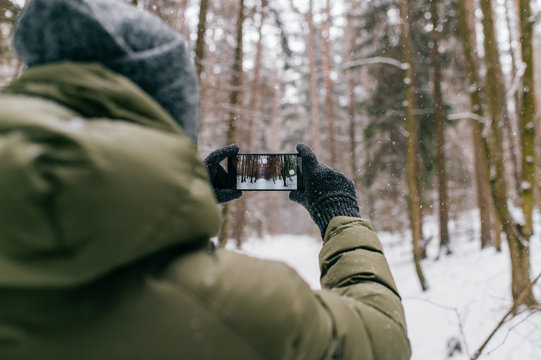 Smartphone in male hands in the winter forest. Sunny winter day.  Man holding mobile smart phone and taking photo, cellphone over blurred snowy forest.