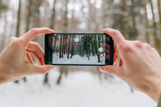 Smartphone in male hands in the winter forest. Sunny winter day.  Man holding mobile smart phone and taking photo, cellphone over blurred snowy forest.