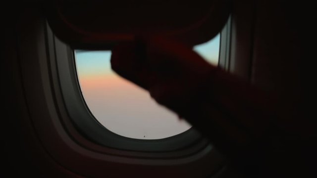 Hand opens airplane window to looks out during air travel. Passenger pov through plane window flight during sunset or sunrise. Close up of porthole of flying airplane at night. Aircraft above clouds.