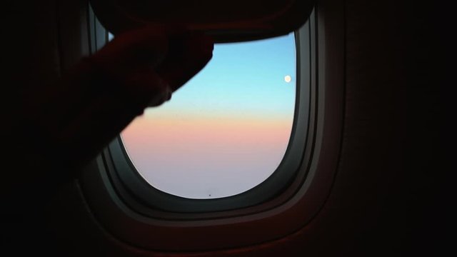Hand opens airplane window to looks out during air travel. Passenger pov through plane window flight during sunset or sunrise. Close up of porthole of flying airplane at night. Aircraft above clouds.