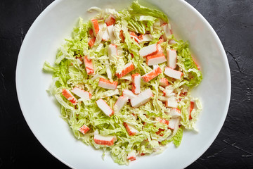 Chopped crab sticks and fresh chinese cabbage leaves in white ceramic bowl on black stone background, top view. Vegetable and imitation crab meat, sea food.  Cooking salad