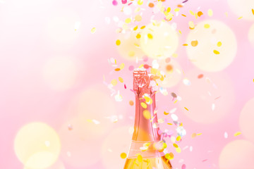 A bottle of sparkling wine with confetti and bokeh in the foreground, on pink background