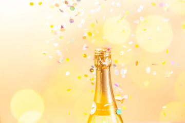 A bottle of sparkling wine with confetti and bokeh in the foreground, on golden background