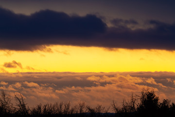 Looking from a vantage point and with a long zoom the clouds below and above create a sandwich with the yellow color of the sun light at sunset.