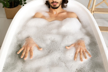 Young man lying in white bathtub filled with hot water and touching foam