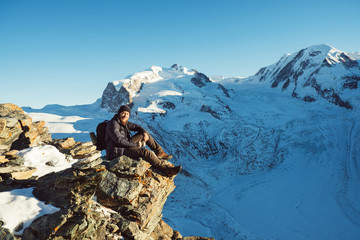 Traveler Man with backpack trekking in mountains, enjoy beautiful Matterhorn view. Explorer man hiking on hills, travel in Swiss Alps, Switzerland. Hiker sitting on rock cliff outdoors on nature.