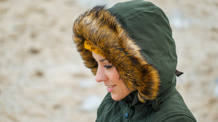 Beautiful winter portrait of young smiling woman in winter snowy scenery. Happy girl in warm clothing with fur hood walk in winter park outdoors. Cheerful woman dressed a green coat and yellow hat.