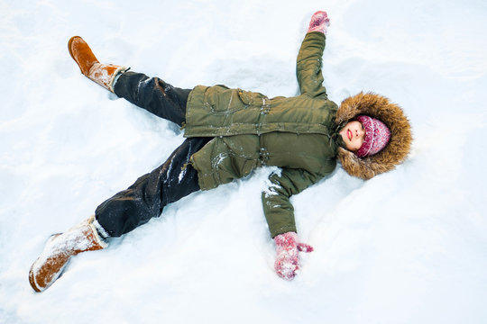 Happy girl laying on snow and making snow-angel. Child have fun and play with snow outdoors on winter nature. Family Christmas holiday background. Walk in park with kids. Kid in winter day.
