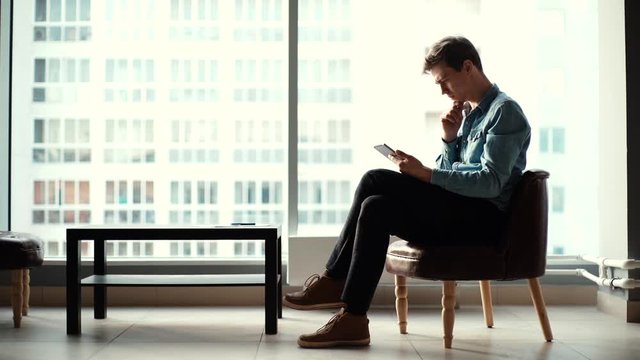 Handsome young business man sitting on a chair and using a tablet in the office against a large window. Shooting in slow motion.
