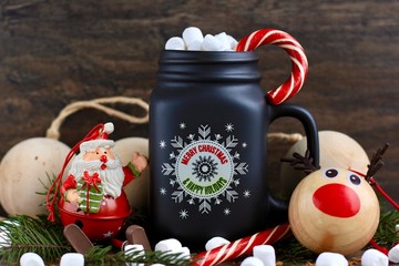 Mug of hot coffee with milk, marshmallow and Lollipop against rustic wooden background with Christmas decorations and lights.