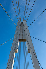 White metal pylon and steel ropes of a suspension pedestrian bridge against a blue sky. They look like a web.