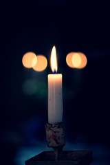 A moody portrait of a burning candle in the dark. From the darkness there appear a few bokeh balls which are actually other lit candles.
