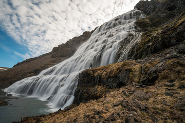 Man stands infront of Dynjandi or Fjallfoss,  big and powerfull waterfall cascade in the westfjords of the Icelandic wilderness during daytime. Waterfalls, traveling and iceland concept.