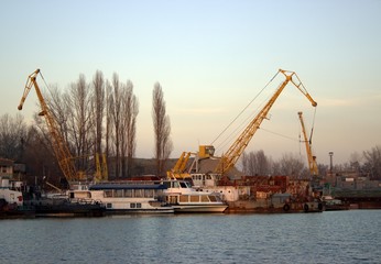 Obraz premium Old yellow cargo cranes in the river port. Nearby are passenger boats.