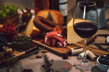 Glass of red wine accompanied by a variety of gourmet ingredients such as a variety of cheeses, cold meats, wine bottles, rustic bread and fruit on a rustic background