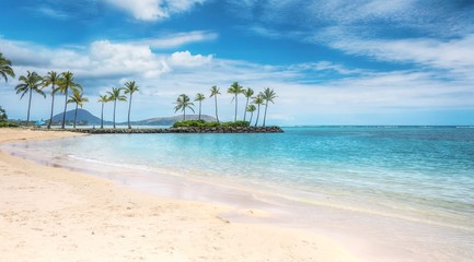 A beautiful beach scene in the Kahala area of Honolulu, with fine white sand, shallow turquoise water, a view of coconut palm trees and Diamond Head in the background.