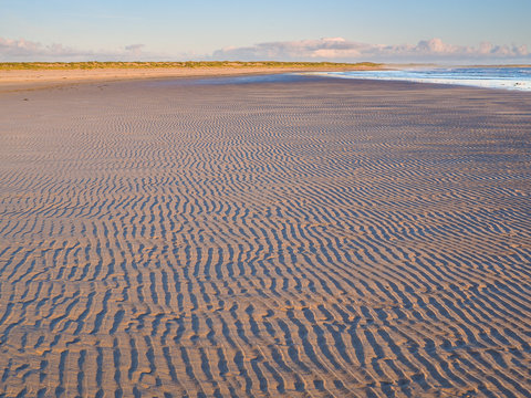 Abstract Rhythmic Patterns and Textures in the Sand Dunes of a Wide, Empty Beach at Sunset or Sunrise with Clear Sky and majestic ruins of Dunstanburgh Castle.