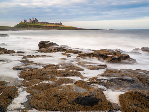 Long exposure of waves crashing against a rocky shore with the ruins of Dunstanburgh Castle on a grassy hill in the background, Northumberland, England.