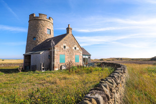 Historic Stone Cottage with a Circular Tower and Dry Stone Wall in a Grassy, Coastal Landscape under a Bright Blue Sky, Scotland Holy Island