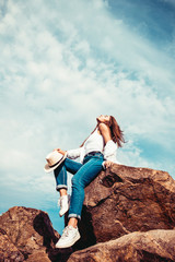 Naklejka premium Beautiful girl in a white shirt, blue jeans and cowboy hat posing on the rock. Outdoor shot. Summer vacation concept.