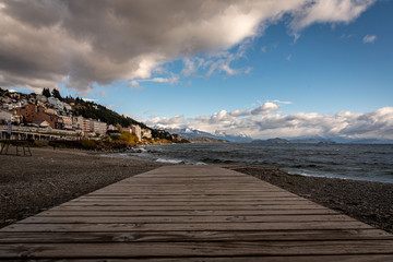 Perspective view of the lake Nahuel Huapi and the cityscape of Bariloche in Argentina, with snowed mountains in the background, seen from wooden deck of stony coast.