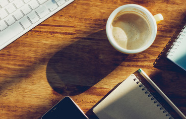 Top view. coffee cup, computer keyboard, mobile phone and notebook on wooden desk with sunlight. Vintage color tone