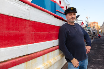 The captain of a very old fishing boat stands in front of his ship. The old wooden boat is painted red and white. The ship is in the harbor in front of a fish hall.