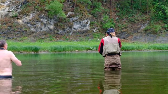 Fly fishing. A photographer with a naked torso waist-deep in water photographs a fisherman. in red clothes. Sports and entertainment on a mountain river.